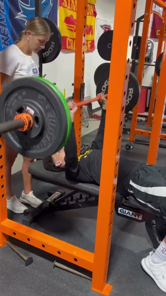 An athlete bench presses a barbell with a spotter in the rack.
